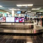 Travelers at baggage claim viewing conveyor belt advertisement in airport terminal