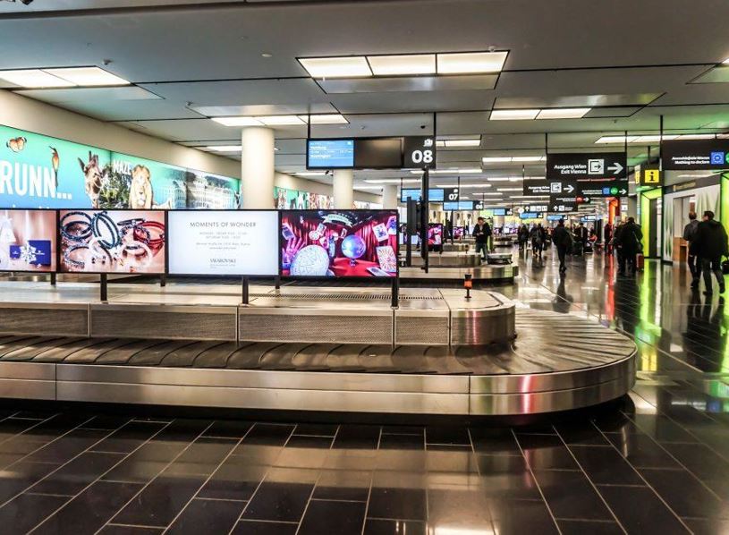 Travelers at baggage claim viewing conveyor belt advertisement in airport terminal