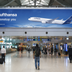 Large Lufthansa airport billboard displayed inside a busy Indian airport terminal with travelers walking below the advertisement.