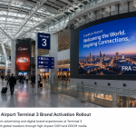 Modern Frankfurt Airport Terminal 3 interior with large digital advertising screens and travelers walking through the terminal.